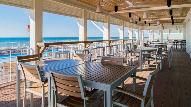Un espace de restauration en plein air avec des tables et des chaises en bois, de grandes fenêtres et une vue sur l’océan.