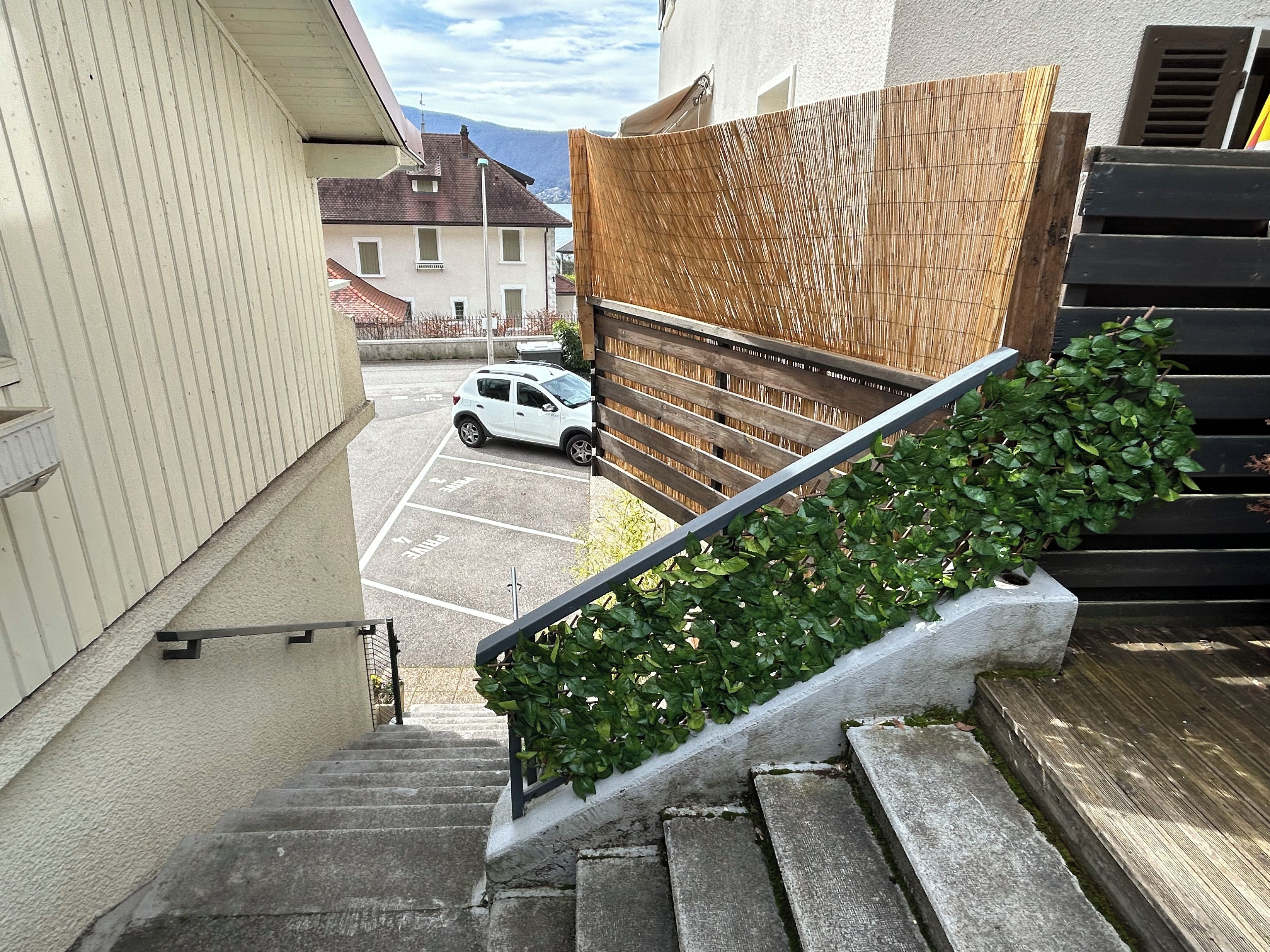 Un barrio residencial con un coche blanco estacionado en una entrada, una escalera que lleva a una terraza de madera, y una vista de una montaña al fondo.