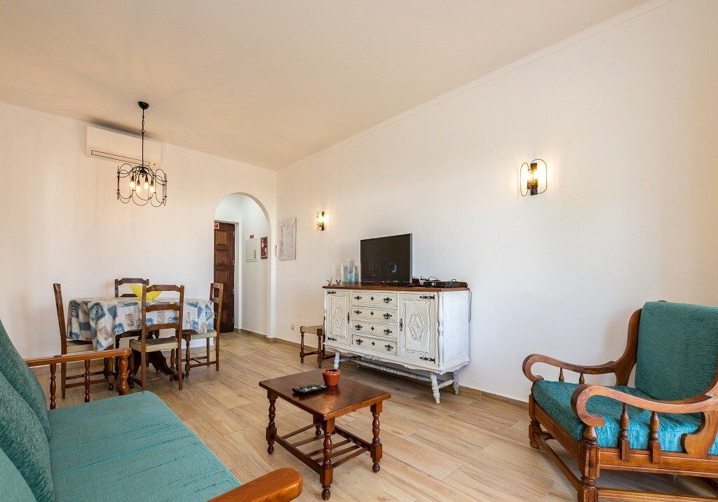 A living room with a sofa, a wooden coffee table, a TV on a white cabinet, and a dining area with chairs and a table.