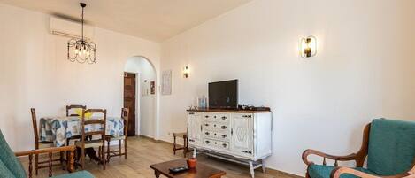 A living room with a sofa, a wooden coffee table, a TV on a white cabinet, and a dining area with chairs and a table.