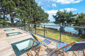 A wooden deck with glass-topped tables and striped beach chairs, overlooking a body of water with boats and trees.