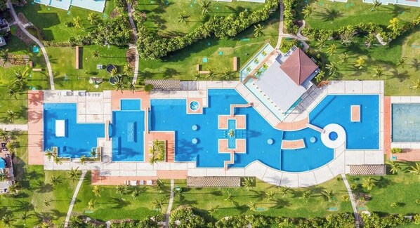 Vue aérienne d’un complexe hôtelier doté de plusieurs piscines, d’un bâtiment central et d’un espace verdoyant environnant.