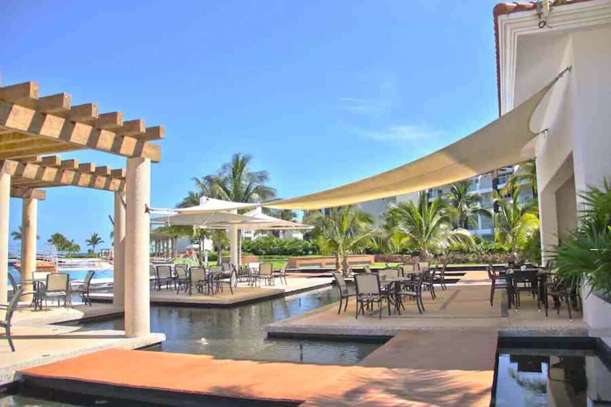 A resort pool area with lounge chairs and umbrellas, surrounded by palm trees and a clear sky.