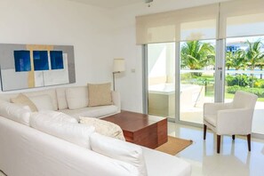 Ein modernes Wohnzimmer mit einem weißen Sofa, einem Holztisch und einem weißen Sessel. Ein großes Fenster bietet Blick auf Palmen und einen Pool.