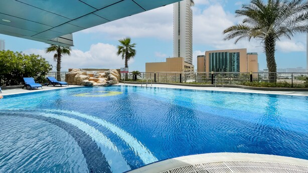Ein Swimmingpool mit klarem, blauem Wasser, umgeben von Liegestühlen und Palmen, mit modernen Gebäuden im Hintergrund.