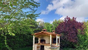Cozinha ao ar livre construída em tijolo, com chaminé, rodeada por vegetação exuberante e árvores.