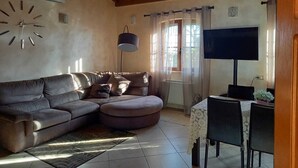 Uma sala de estar com sofá seccional, televisão, mesa com cadeiras e relógio na parede.