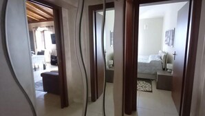 Um corredor com design moderno que leva a um quarto com cama, mesa de cabeceira e luminária. Uma área de jantar é visível ao fundo.