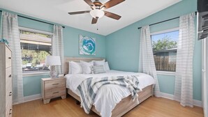 Une chambre à coucher avec un lit, une table de chevet, un ventilateur de plafond et une fenêtre avec des rideaux.