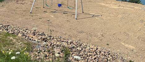 A playground with swings and a dirt path leading to a body of water.