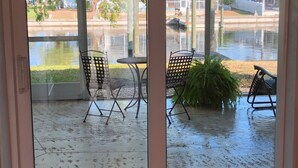 A view through a glass door of a patio area with a table and chairs, a small water feature, and a view of a park and buildings in the background. - Lovely House on the canal with private dock, large fenced yard & pet friendly (Apollo Beach)