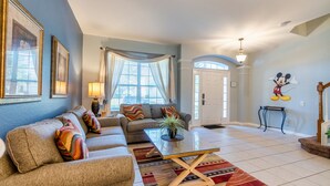 A living room with a sofa, a coffee table, and a large window with curtains. - Lakefront Windsor Hills - 6 Bed 4 Bath Home with Custom Mickey Mouse Heated Pool (Kissimmee)