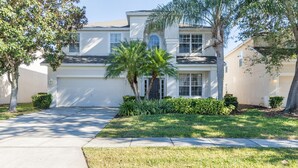 A two-story house with a garage, a well-maintained lawn, and mature trees. - Lakefront Windsor Hills - 6 Bed 4 Bath Home with Custom Mickey Mouse Heated Pool (Kissimmee)