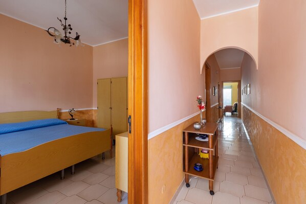 A bedroom with a bed, a wooden wardrobe, a nightstand, a shelf with various items, and a hallway leading to another room.