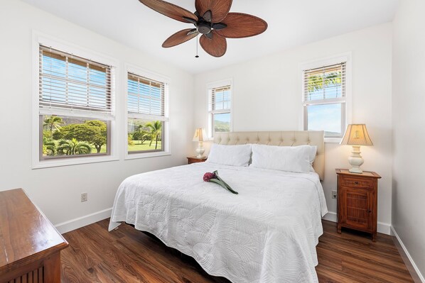 A bedroom with a bed, wooden floor, ceiling fan, two windows with blinds, a nightstand with a lamp, and a view of greenery outside.