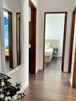 Corredor que dá para um quarto com cama, mesa de cabeceira e criado-mudo.