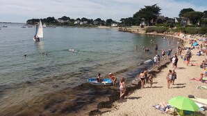 A beach with clear water, people swimming and sunbathing, and a sailboat in the distance.