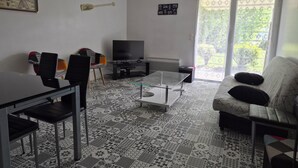 A living room with a TV, a dining table, chairs, a patterned floor, and a sofa with pillows. - Appartement Avec Terrasses, Équipements Entièrement Neufs (Crozon)