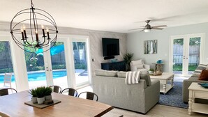 Ein geräumiges Wohnzimmer mit grauem Sofa, einem Esstisch mit Stühlen, einem Deckenventilator und Blick auf einen Swimmingpool durch große Glasüren.