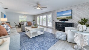 Ein geräumiges Wohnzimmer mit Deckenventilator, großem Flachbildfernseher an der Wand und Blick auf den Strand durch die Fenster.