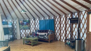 A cozy room with a wooden floor, a sofa with a bear-themed blanket, a small table, and a metal heater. - Pet-Friendly Yurt Sleeps 12 with Pool Access (Granville)