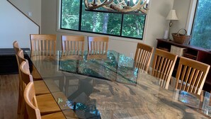 Een eethoek met een tafel met glazen blad, omringd door houten stoelen, een kroonluchter met gewei en uitzicht op bomen door grote ramen.