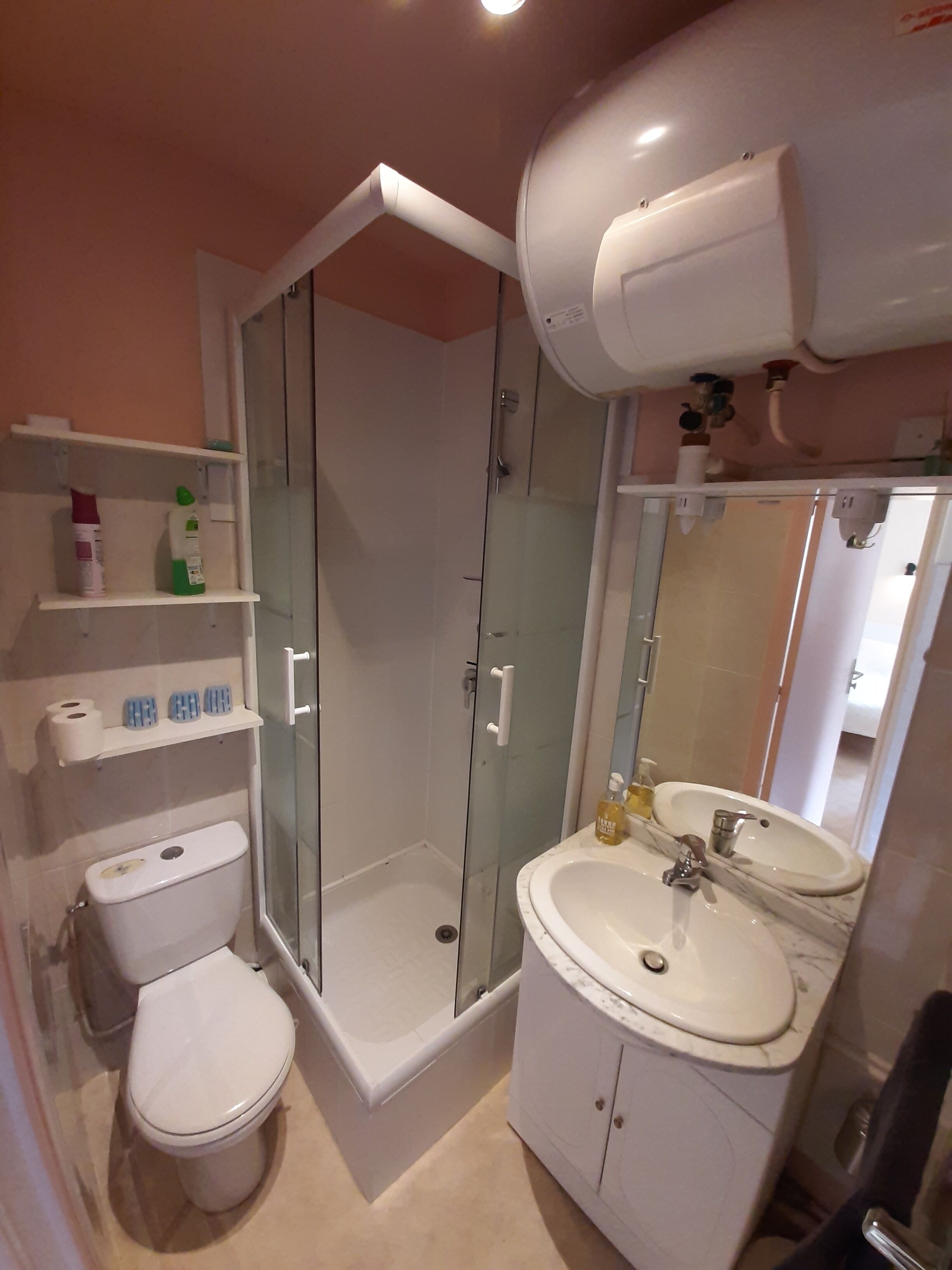 Un baño con inodoro, ducha con cerramiento de vidrio, lavamanos con espejo y repisas con artículos de aseo.