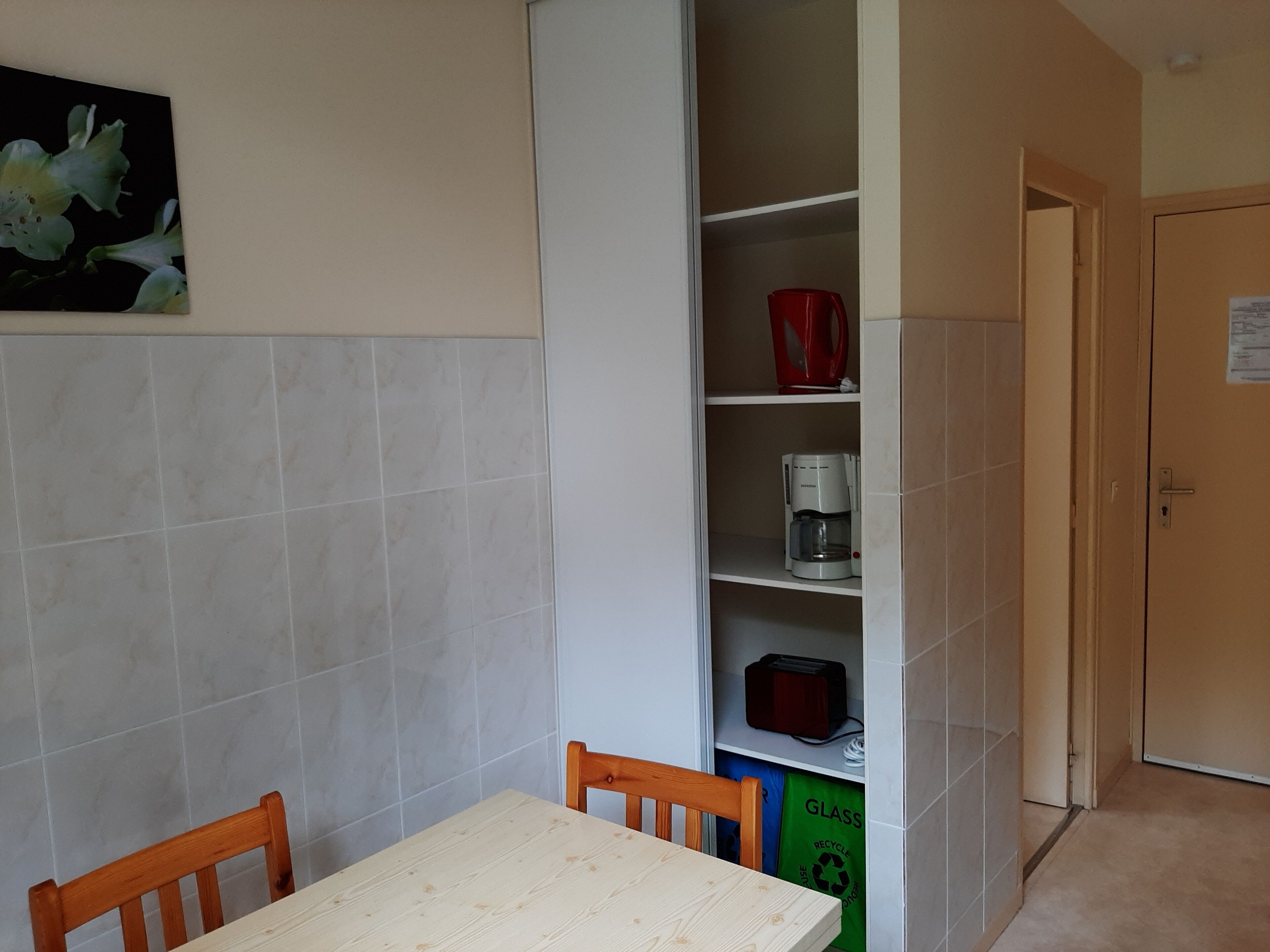 Une cuisine avec un mur recouvert de carreaux blancs, une table en bois et des chaises. On y voit une étagère avec une tasse rouge, une machine à café et un sac de recyclage. Une porte est visible au fond.