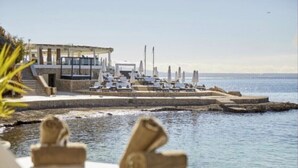 A seaside resort with a pool area, lounge chairs, and a building with a terrace overlooking the water. - Apartamento-casa de Diseño con Entrada Privada y Pequeña Piscina (ses Illetes)