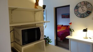 A compact room with a white microwave, a clock, a small plant, and a bed with a colorful blanket.