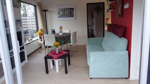 A living room with a light blue sofa, a dining table with a red and green tablecloth, and a white chair. There is a framed artwork on the wall and a window with curtains.