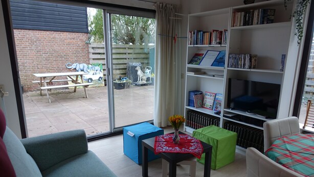 A living room with a sofa, a small table with a vase of flowers, a bookshelf, and a sliding glass door leading to an outdoor area with a picnic table and a dog.