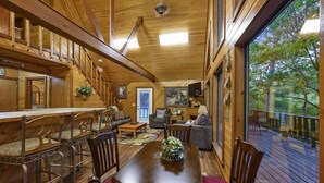 Interior de una cabaña de madera con comedor, sala de estar y un balcón con vistas al bosque.