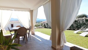 An outdoor patio with a table and chairs, white curtains, and a view of a garden and buildings in the background. - Villa a 600m dal Mare (Budoni)