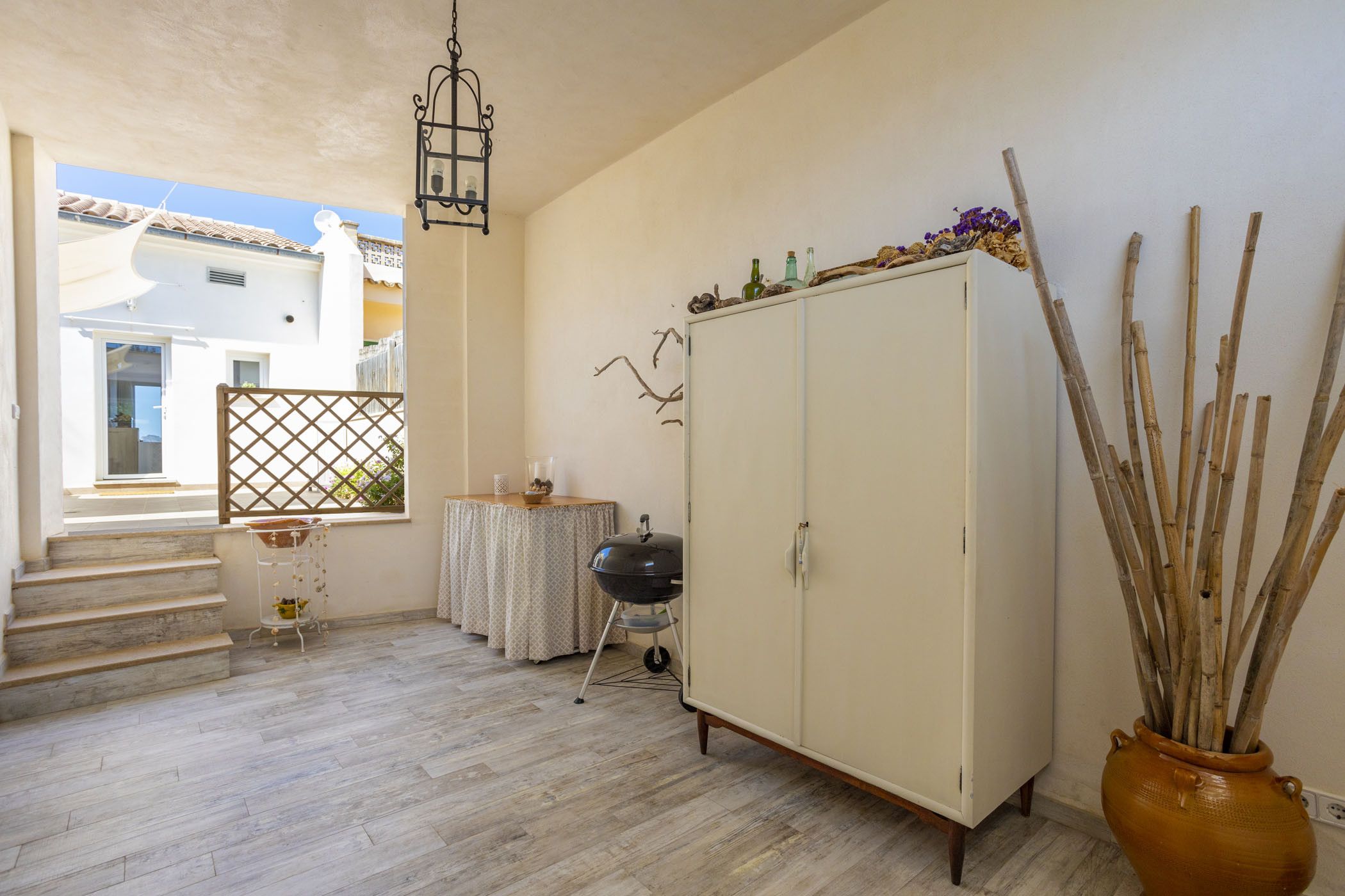 A room with a wooden floor, a white wardrobe, a small round table with a white tablecloth, a hanging light fixture, and a window with a view of a building outside.