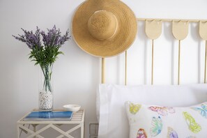 A bed with white pillows, a wooden headboard, a straw hat, a vase with flowers, and a small table with a book and bowl.