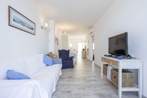 A living room with a white sofa, a blue armchair, a TV on a white stand, and a hallway leading to a dining area.