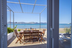 A covered outdoor seating area with a table and chairs, overlooking a beach and the sea.
