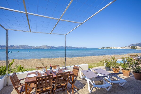 Ein überdachter Außenbereich mit Tisch und Stühlen, mit Blick auf einen Strand und das Meer.
