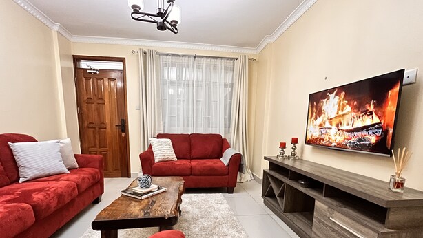 A living room with a red sofa, a wooden coffee table, and a flat-screen TV displaying a fire scene. - Neema Executive Suites Near Junction Mall 5A (Nairobi)
