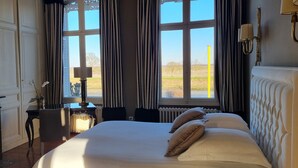 A hotel room with a large bed, a fireplace, a desk with a lamp, and large windows with curtains.
