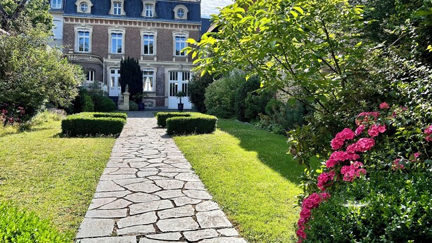 A stone pathway leading to a large, historic building with a symmetrical facade, surrounded by well-maintained hedges and flowering plants.