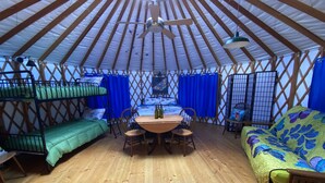 A yurt with a wooden floor, a central table with chairs, bunk beds, and a colorful sofa.