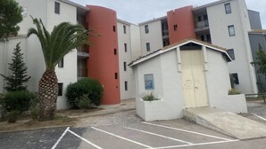 Un edificio residencial de varios pisos con una estructura cilíndrica roja en el centro, un garaje pequeño adosado y una palmera en primer plano.