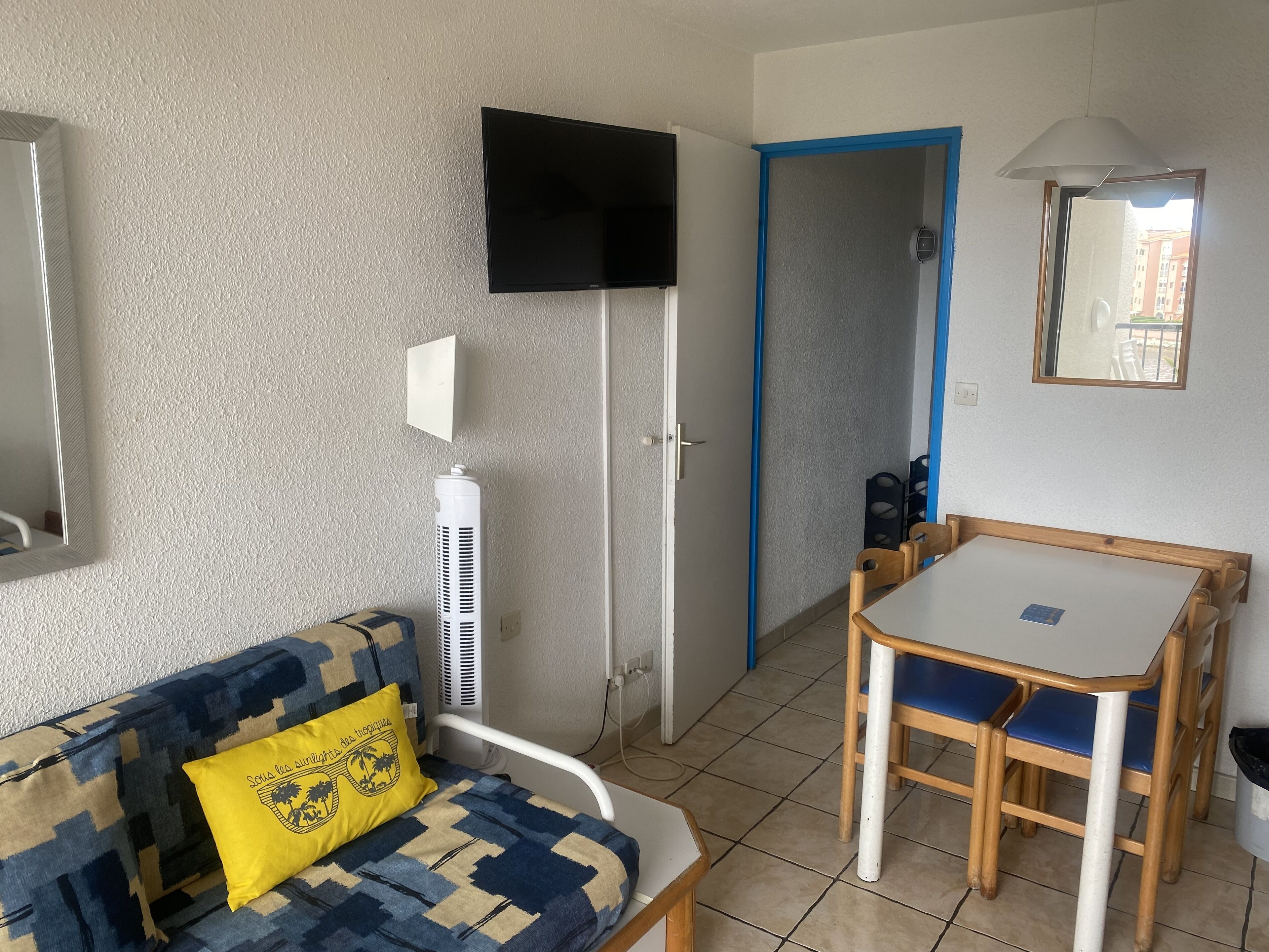 A compact living space with a sofa, a small table with chairs, a TV, and a door leading to another room.