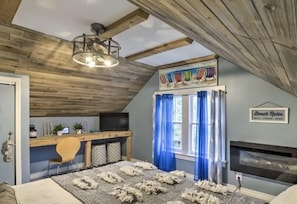 A cozy attic bedroom with a wooden ceiling, a desk with a chair, a large window with blue curtains, and a fireplace.