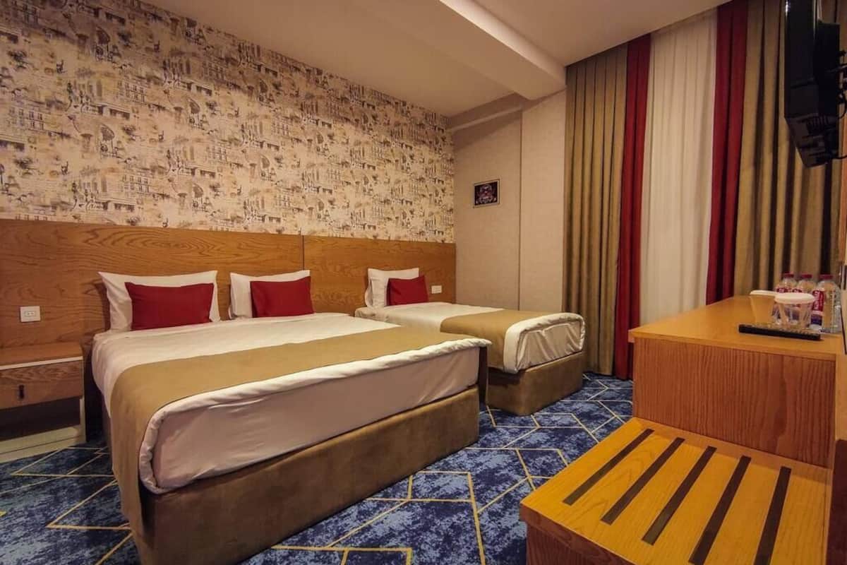 Ein Hotelzimmer mit zwei Betten, einem hölzernen Kopfteil, einem blau gemusterten Teppich, einem Nachttisch aus Holz und einer Wand mit einem Stadtlandschaftsmural.