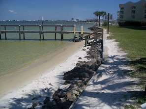 A beach with a wooden walkway, a stone wall, and a view of the water and distant buildings.