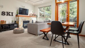 A modern living room with a fireplace, a sofa, a dining table with chairs, and a television mounted on the wall. - 2BR | Symphony | Dog Friendly | Hot Tub | Private Balconies | Village Location | Parking (Whistler)