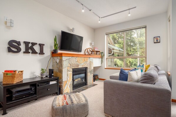 A living room with a stone fireplace, a flat-screen TV, a sofa, and a large window with a view of trees. - 2BR | Symphony | Dog Friendly | Hot Tub | Private Balconies | Village Location | Parking (Whistler)